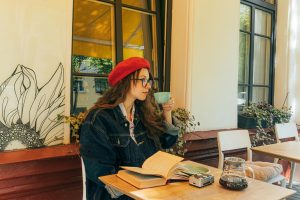Stylish woman in red beret sipping coffee at a cafe with an open book and denim jacket.