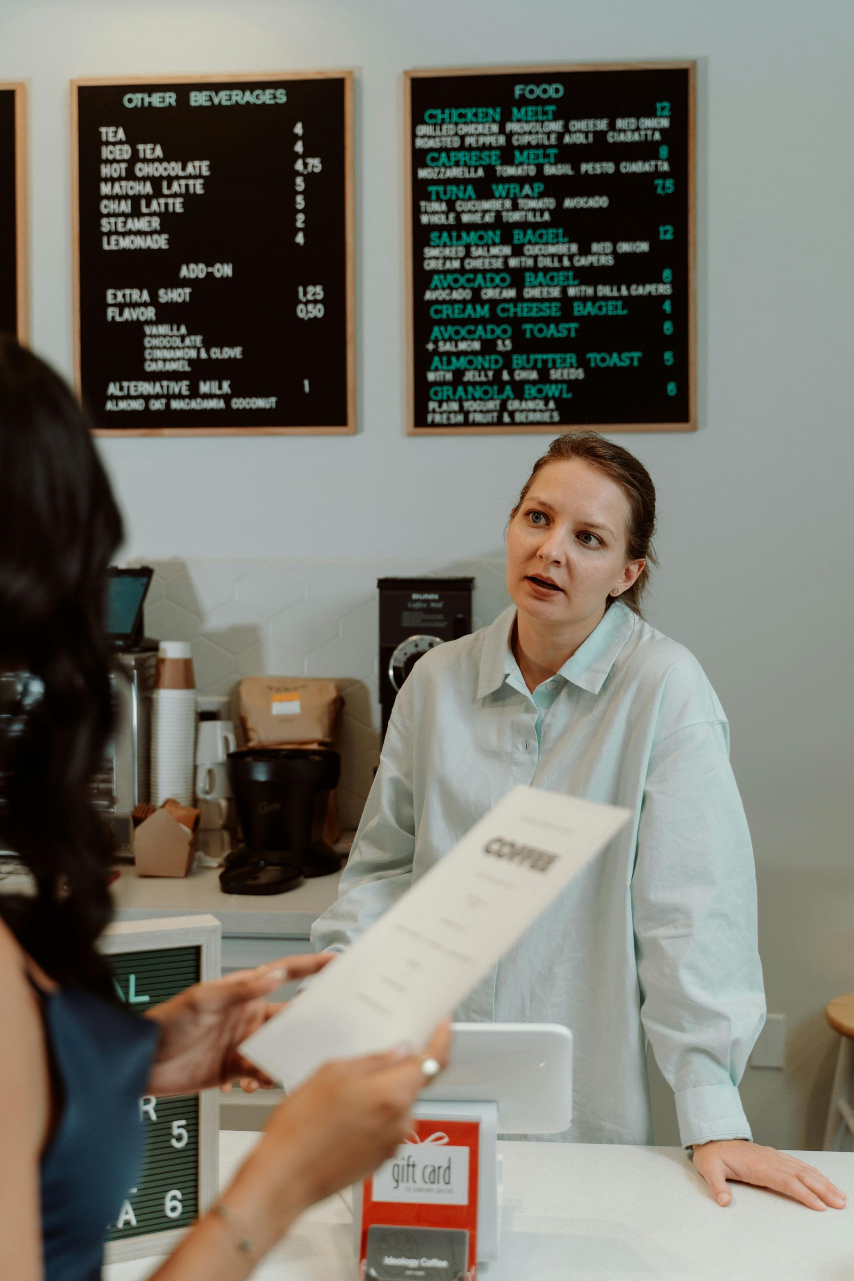 A woman at a café counter engaging with a customer holding a menu. Beverage and food menu visible.